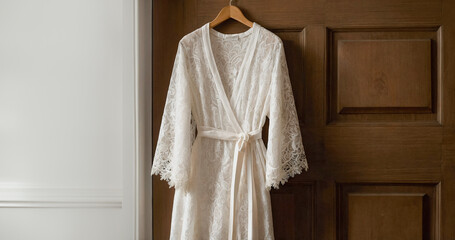 White lace dressing gown with long sleeves and neckline hanging in bedroom. Luxury elegant dress with lace