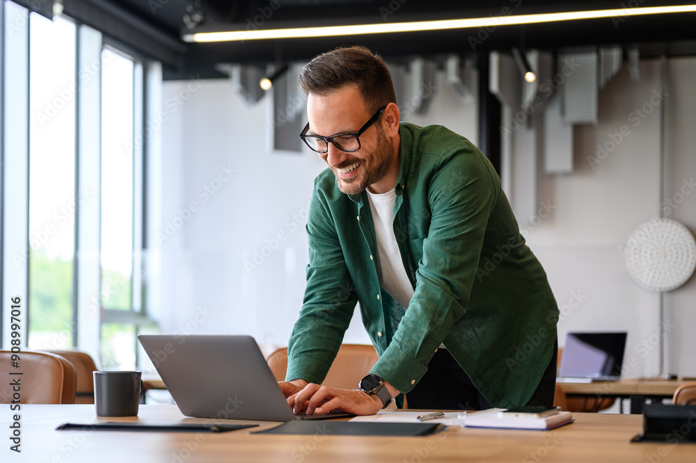 Wall mural young businessman in glasses smiling and typing over laptop computer while standing at desk in offic