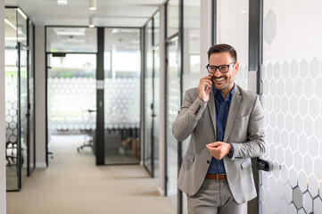 Fototapeta premium Successful male entrepreneur smiling and talking on mobile phone while standing by glass door in office