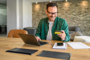 Male accountant in glasses paying bills with credit card over laptop while sitting at his workplace