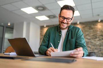 Low angle view of project manager writing on paper while doing online research over laptop at desk