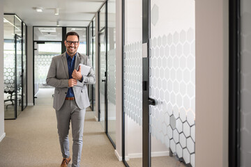 Portrait of smiling handsome CEO in glasses holding computer and walking happily in office corridor