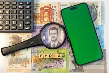 Bashar al-Assad portrait on syrian banknotes is focused in magnifying glass and smartphone with green blank screen for text