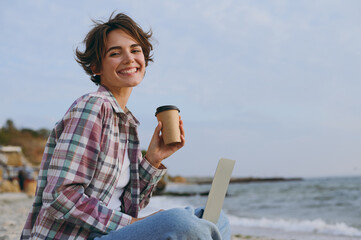 Side view young IT woman wears shirt casual clothes hold use work on laptop pc computer drink...
