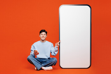 Full body young fun man wears blue sweatshirt casual clothes point on big huge blank screen mobile cell phone with area use smartphone isolated on plain red orange color background. Lifestyle concept.