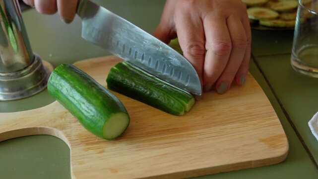 Slices a fresh cucumber on a wooden cutting board, using a sharp knife. The scene is set in a kitchen