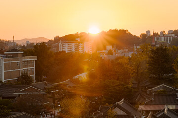 sunset drapes over Jeonju Hanok Village, silhouetting traditional houses against a twilight sky, as a bustling street comes alive under the warm, atmospheric light.