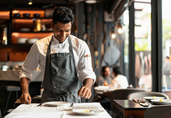 Fototapeta premium A handsome young Indian man wearing a chef attire standing at the head of an empty table