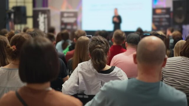 Speaker - young adult woman - speaking to a large audience. Eager audience at a creative industry event, speaker presenting concepts, slightly out of focus. Young men and women in casual wear
