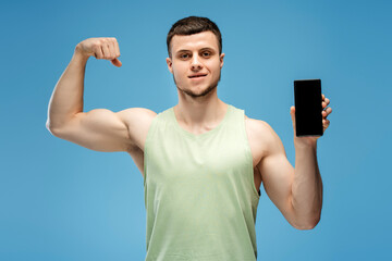 Athletic man showing muscles and holding smartphone with blank screen, isolated on blue background