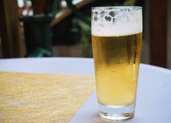 Glas Bier steht auf Tisch mit weißer Stofftischdecke und goldener Papierserviette mit Muster bei Feier in Garten am Abend im Sommer