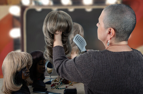 Woman with a shaved head combs her ash blonde wig, with a variety of wigs on display on her vanity.