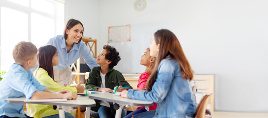 Happy diverse kids sitting at desk in classroom and talking with teacher, studying during lesson,...