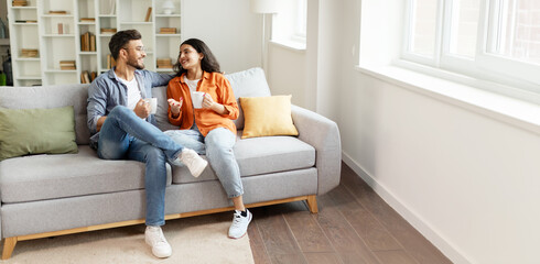 Happy indian couple relaxing and drinking coffee, sitting on couch at home, talking , free space