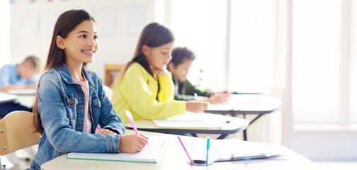 Smiling cute European schoolgirl in casual attentively listening to teacher while sitting at her...