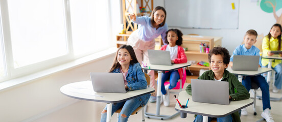 School children using laptops, female teacher pointing at blackboard during exam test at computer science class. Modern education