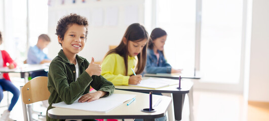 Portrait of smiling Latin schoolboy showing thumbs up and smiling, sitting at desk in his classroom...