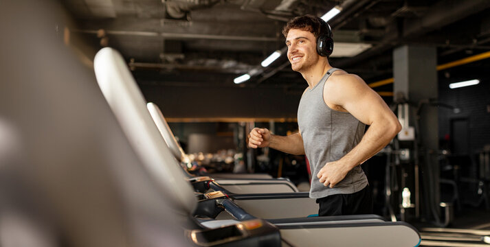Handsome man in wireless headphones jogging on treadmill at gym and listening music while running, enjoying workout, copy space