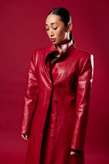 Fototapeta premium Asian fashion beauty in red leather coat posing against red background with hands on hips