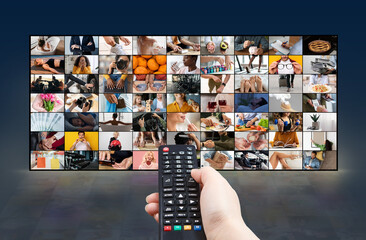 A hand holds a black remote control in front of a large screen displaying numerous images. The...