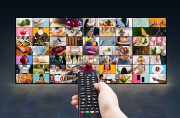 A persons hand holds a black remote control in front of a large television screen. The television screen is displaying a grid of numerous colorful images, depicting various scenes and activities