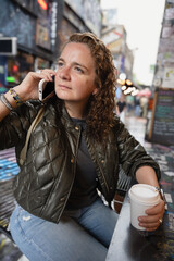 Girl drinking coffee from a disposable cup and talking on the phone in a cafe with a blurred urban background.