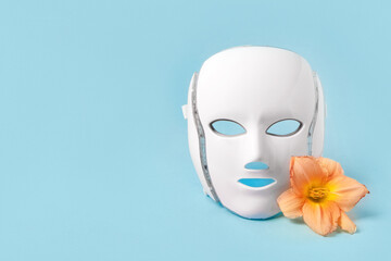 Cosmetic LED facial mask regenerative treatment on blue background with flowers lilies. Health and...