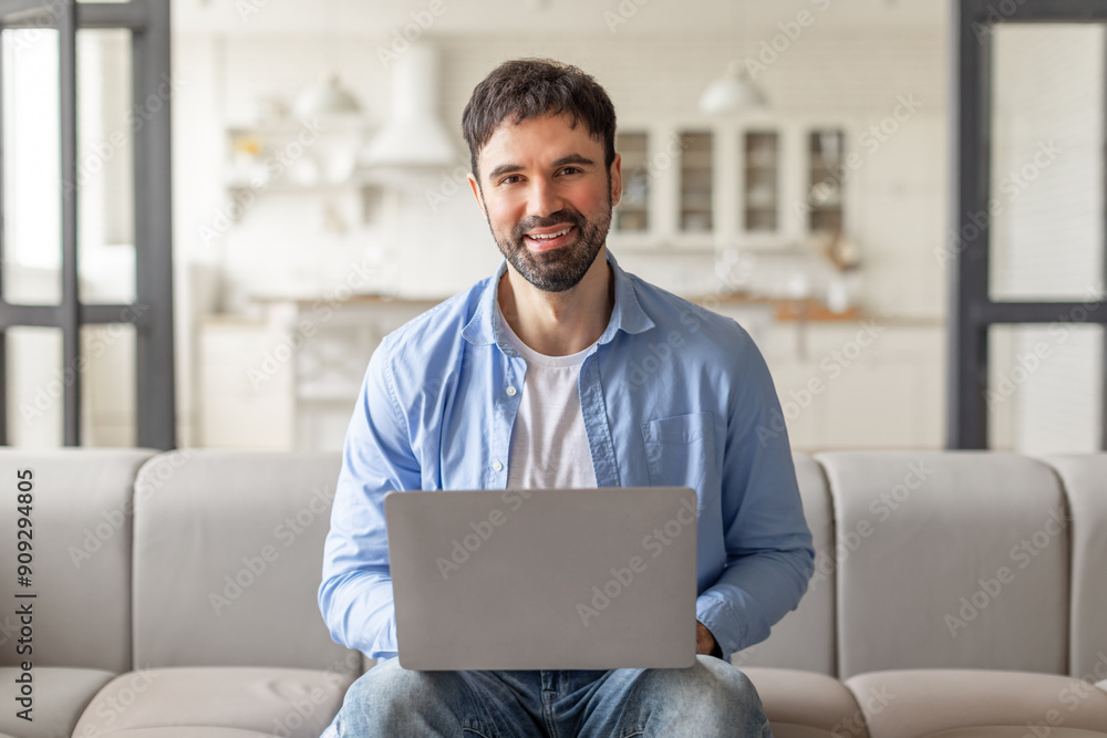 Poster A man is sitting on a couch in a modern home, using a laptop computer. He is smiling and looking directly at the camera. The background is blurred and shows a light-colored living room