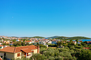 Obraz premium View on coastal town with red roofed houses and lush green hills under clear blue sky. Touristic Mediterranean town near Adriatic sea for summer holidays