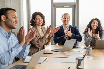 A group of diverse colleagues is sitting around a table in a modern office space. They are clapping and smiling, clearly pleased with the outcome of a successful meeting