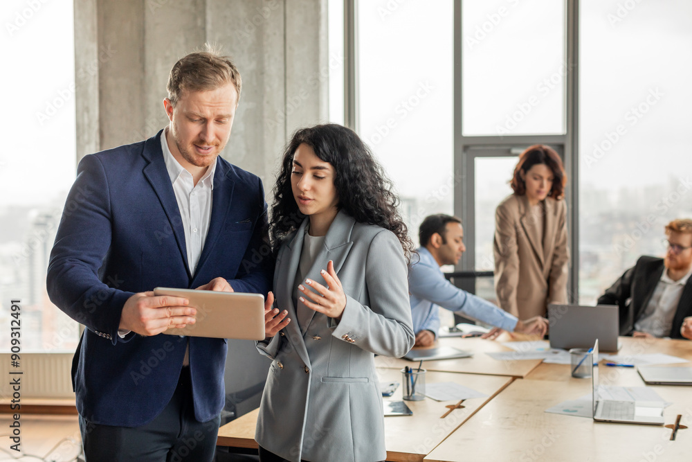 Poster A man and a woman in business attire are discussing a project while looking at a tablet in a modern office setting. They are standing in the foreground with other colleagues working in the background