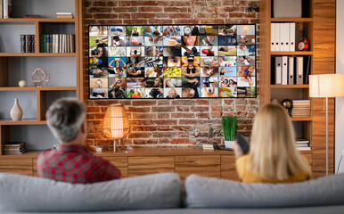 A couple sits on a couch in a living room, facing a large television screen displaying a mosaic of images. The room features a brick wall and wood accents, suggesting a comfortable and cozy atmosphere