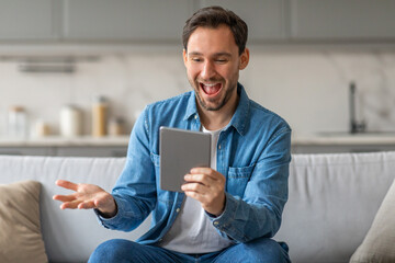 A man sits on a couch in a living room, looking excitedly at a tablet in his hands. He smiles and looks surprised as he reads the screen.