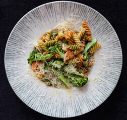 Fusilli pasta in creamy sauce with salmon and broccoli. Tricolor pasta in a white plate on a black background.
