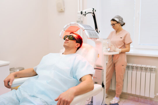 Trichologist female adjusting innovation device for photodynamic therapy procedure after hair transplant of male patient with hairloss problem at medical clinic. Concept of hair transplant procedure.