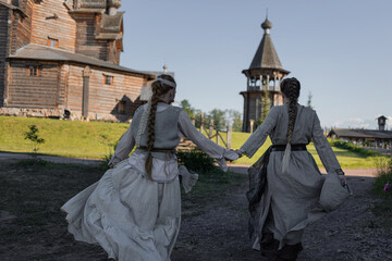 Young women in folk costume run forward holding hands in a village among wooden architecture,...