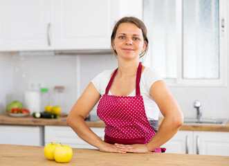 Young woman wearing apron stands at light kitchen, putting your hands on the table