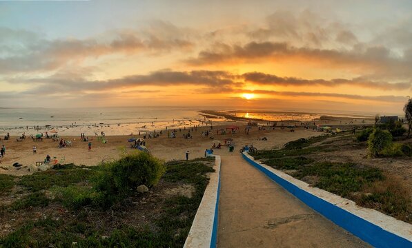 Sidi bouzid, Morocco - 4 August 2024 : A picturesque sunset over a walkways to the beach featuring unicolored umbrellas and chairs for rent on the sand. People swim and walk along the shoreline