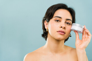 Serious-looking young woman holds a moisturizer jar near her face, presenting a beauty product