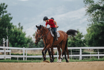 Young girl riding a horse during a training session at an outdoor equestrian facility with scenic mountain and tree background.