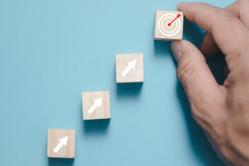 Close up of man hand arranging wood block stacking as step stair on blue background, putting down the cube with target icon on the top . Growth, success, strategy concept