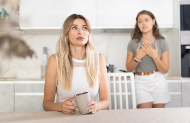 Upset mother sitting at the kitchen-table with her back to her young daughter disputing with her