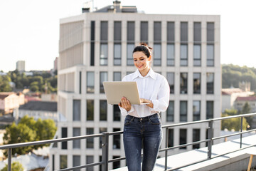 Smiling young woman using laptop on rooftop with modern city skyline in background. Business professional enjoying outdoor workspace with urban scenery.