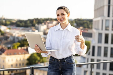 Smiling woman holding laptop and giving thumbs up on rooftop with cityscape background. Woman wearing casual white shirt and jeans, representing modern remote work lifestyle.