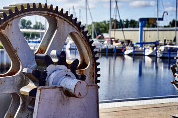 Rusty gear contrasts with a busy marina on a sunny day, adding industrial charm. Blurred focus...