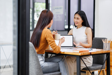 Creative team working on a project in loft office. Two happy asian women analyzing documents discussing marketing plan. Laptop and paperwork on the table