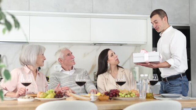 Adult son hands gift, box tied with ribbon, to elderly mom during feast, family holiday dinner. Senior spouses celebrate event in cozy family atmosphere in company of younger couple