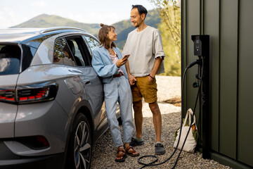 Portrait of young lovely couple standing together near a house and electric car outdoors. Concept...