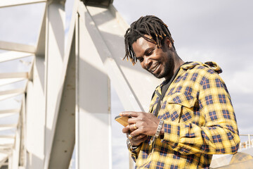 smiling young black man looking at his mobile phone with a modern metallic structure in the background, lifestyle concept, copy space for text