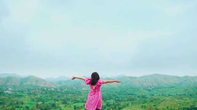 Pretty Indian girl in pink dress raise hands while standing against green hills at Rayta in Udaipur, Rajasthan, India. Tourist enjoying holidays. Cloudy monsoon weather at hill station.